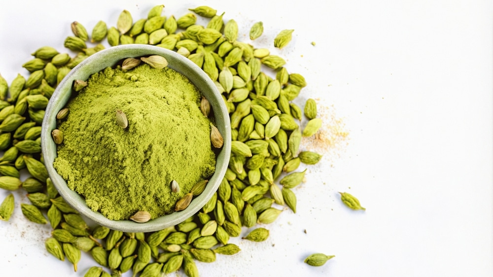 A close-up of green cardamom powder in a bowl surrounded by whole cardamoms.