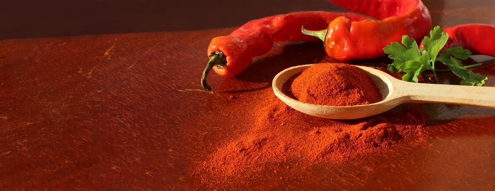 Red paprika seasoning in wooden spoon side view next to peppercorns on wooden red cracked background