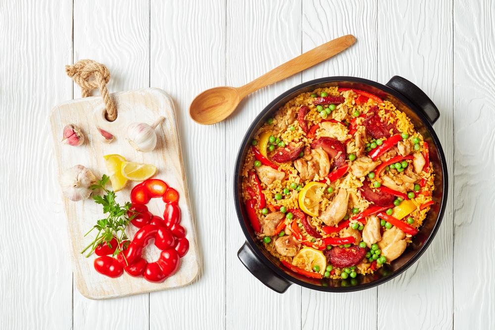 delicious spanish chicken paella with valencian bomba rice, chicken thigh meat, chorizo sausages, vegetables and spices in a black dish on a white wooden table